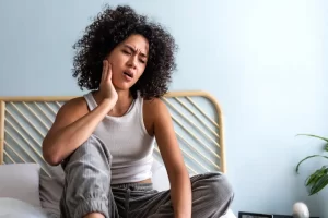 lady sitting on bed holding jaw in pain, TMJ pain